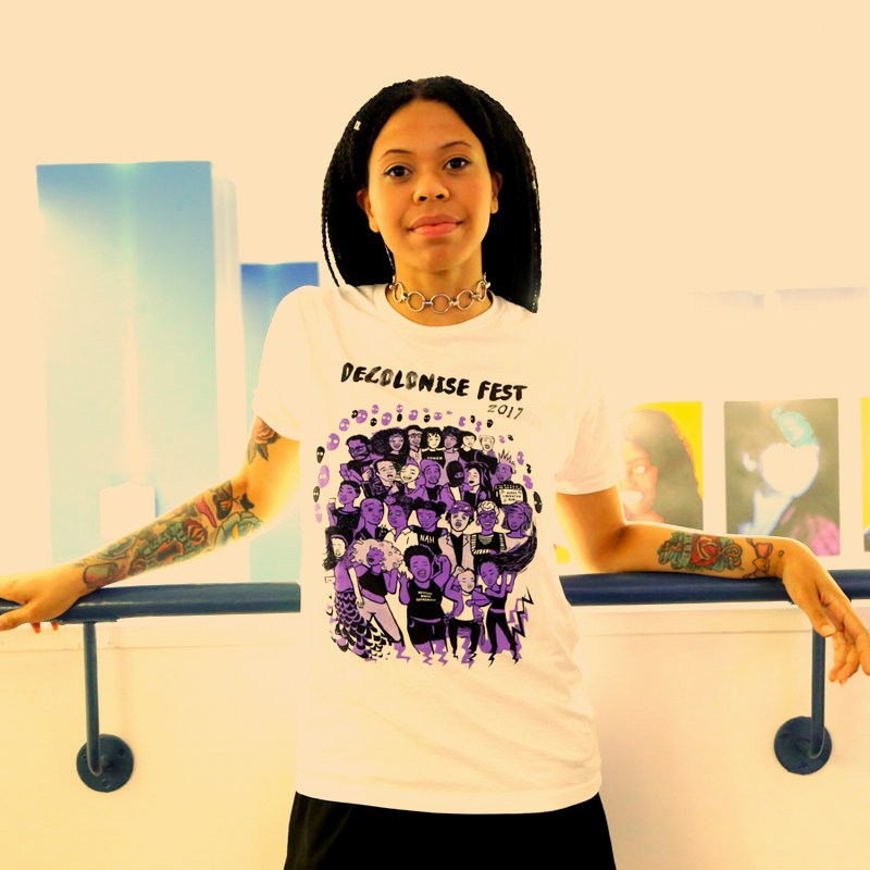Woman standing against a white wall modelling a white Decolonise Fest T-shirt with a purple Decolonise Fest print on it