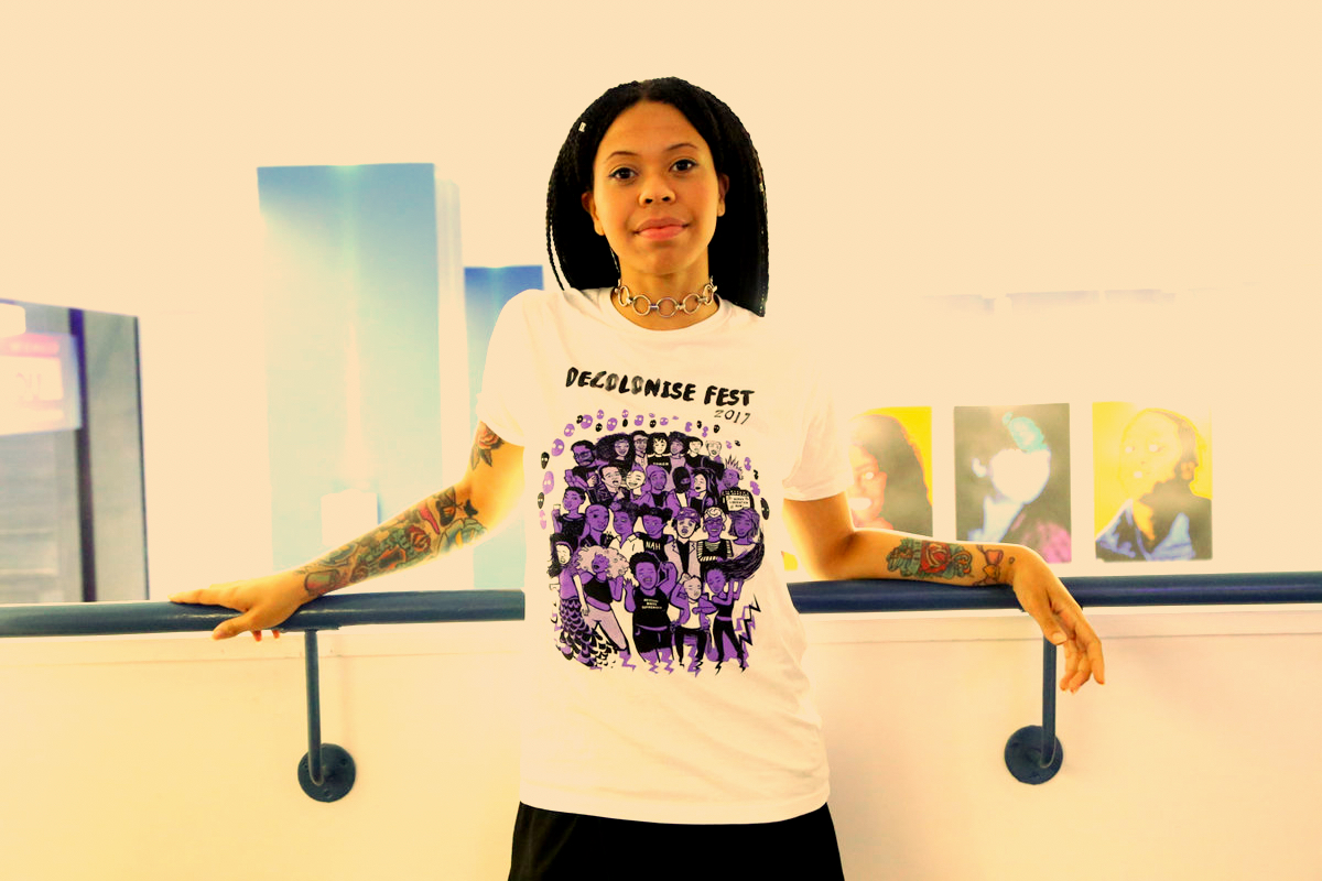 Woman standing against a white wall modelling a white Decolonise Fest T-shirt with a purple Decolonise Fest print on it