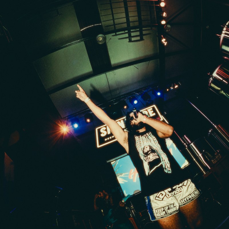 Hyphen performing with his fist raised up. Long hair, black tshirt, white shorts with Hyphen logo